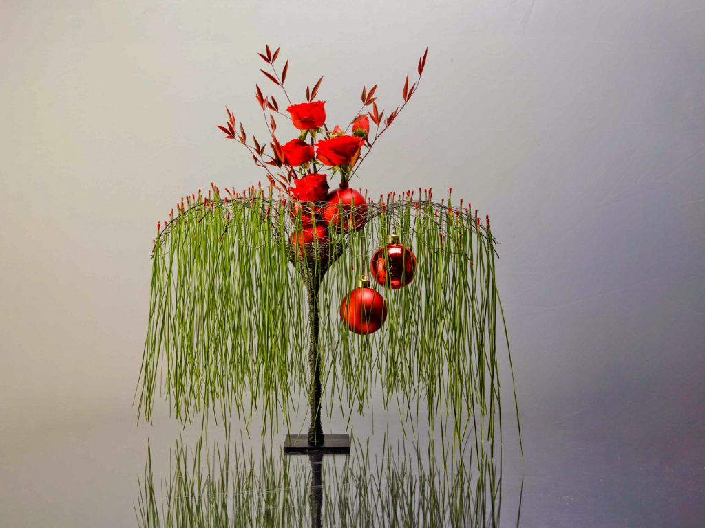 Beautiful round steel wire structure decorated with green pine needles and Christmas balls hanging down. Red ilex  and red roses decorate the top part of the composition.